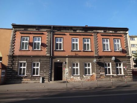 Widok ma fasadę budynku przy ulicy Oświęcimskiej 20 w Chrzanowie przed modernizacją. Fasada w kolorze czerwono-szarej z widocznymi  uszkodzeniami w postaci oderwanego tynku przy poziomie gruntu.