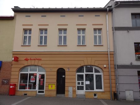 Widok na ścianę frontową przy ulicy Rynek 10 w Chrzanowie. Fasada w kolorze beżowo-pomarańczowym. Na wprost wejście główne do budynku, po prawej stronie wejście do lokalu użytkowego, po lewej stronie wejście do Poczty Polskiej.